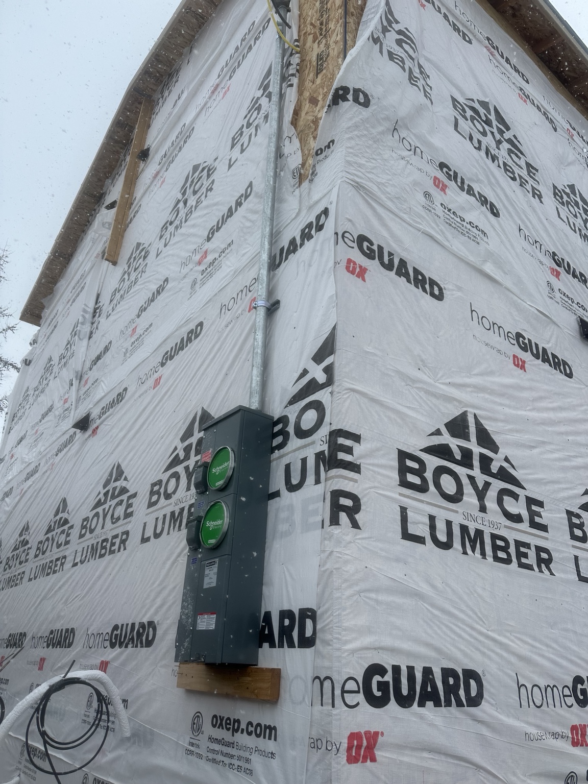 Exterior meter enclosure with two green socket covers and vertical conduit mounted on a house wrapped in building membrane during construction