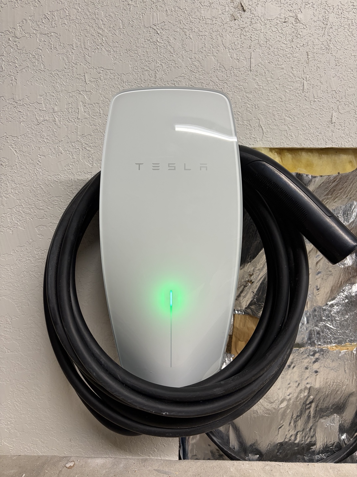 White Tesla wall connector with a green indicator light and coiled black charging cable mounted beside an unfinished wall opening