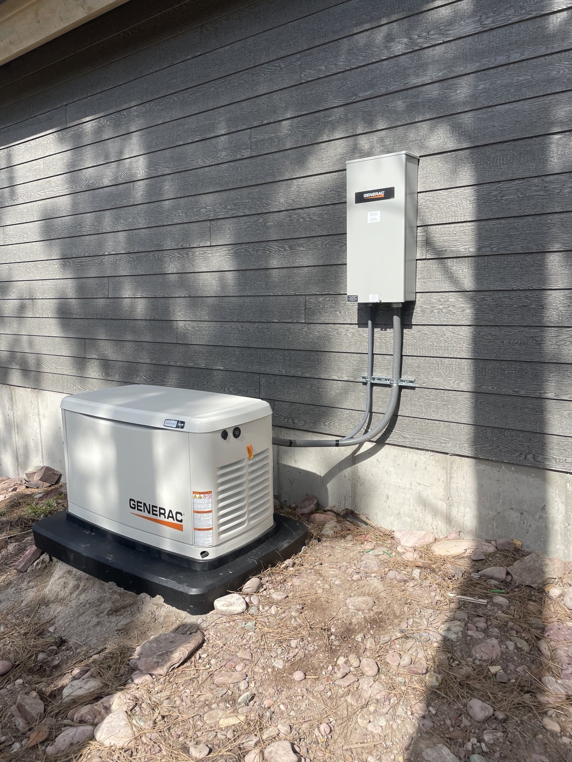 Generac standby generator on a black base beside dark siding, connected by conduit to a wall-mounted exterior enclosure