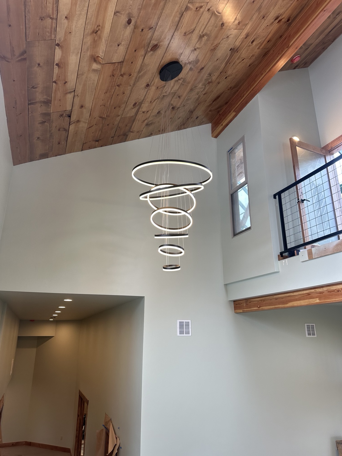 Tiered illuminated ring chandelier hanging in a tall stairwell under a wood ceiling
